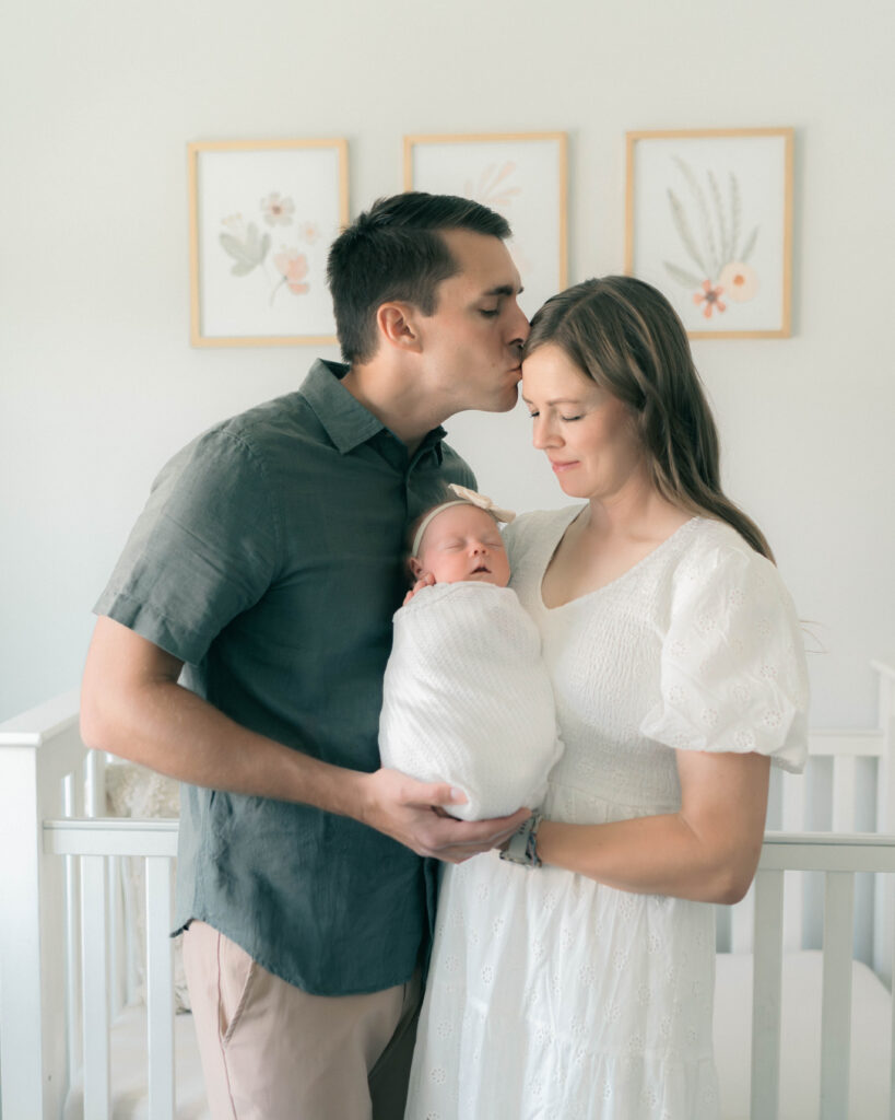 Mother and father holding newborn during an in-home session in Utah, wearing her own light, elegant dress. Captured in soft natural light by Utah newborn photographer.